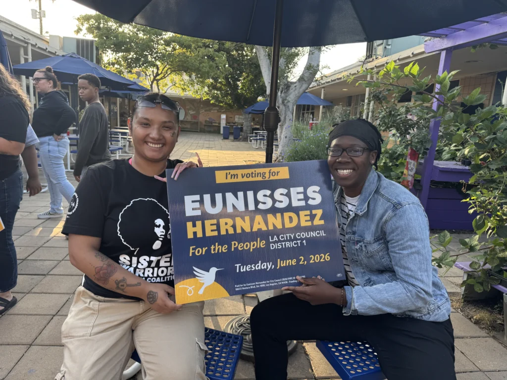 Two women sit at a patio table holding an I'm Voting for Eunisses Hernandez sign.