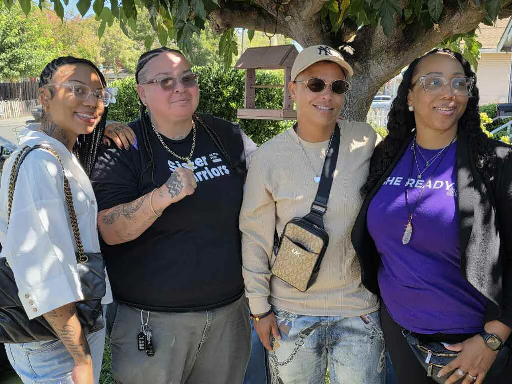 Four people stand arm in arm outside, one in a Sister Warriors shirt and one in purple.